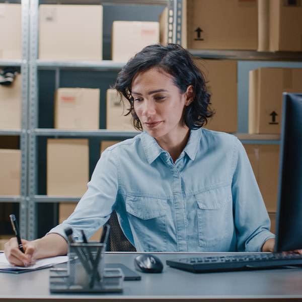 A Woman In A Blue Shirt Manages Kingfisher Grease Fittings And Stock At A Busy Warehouse Desk, Ensuring Global Order Precision.