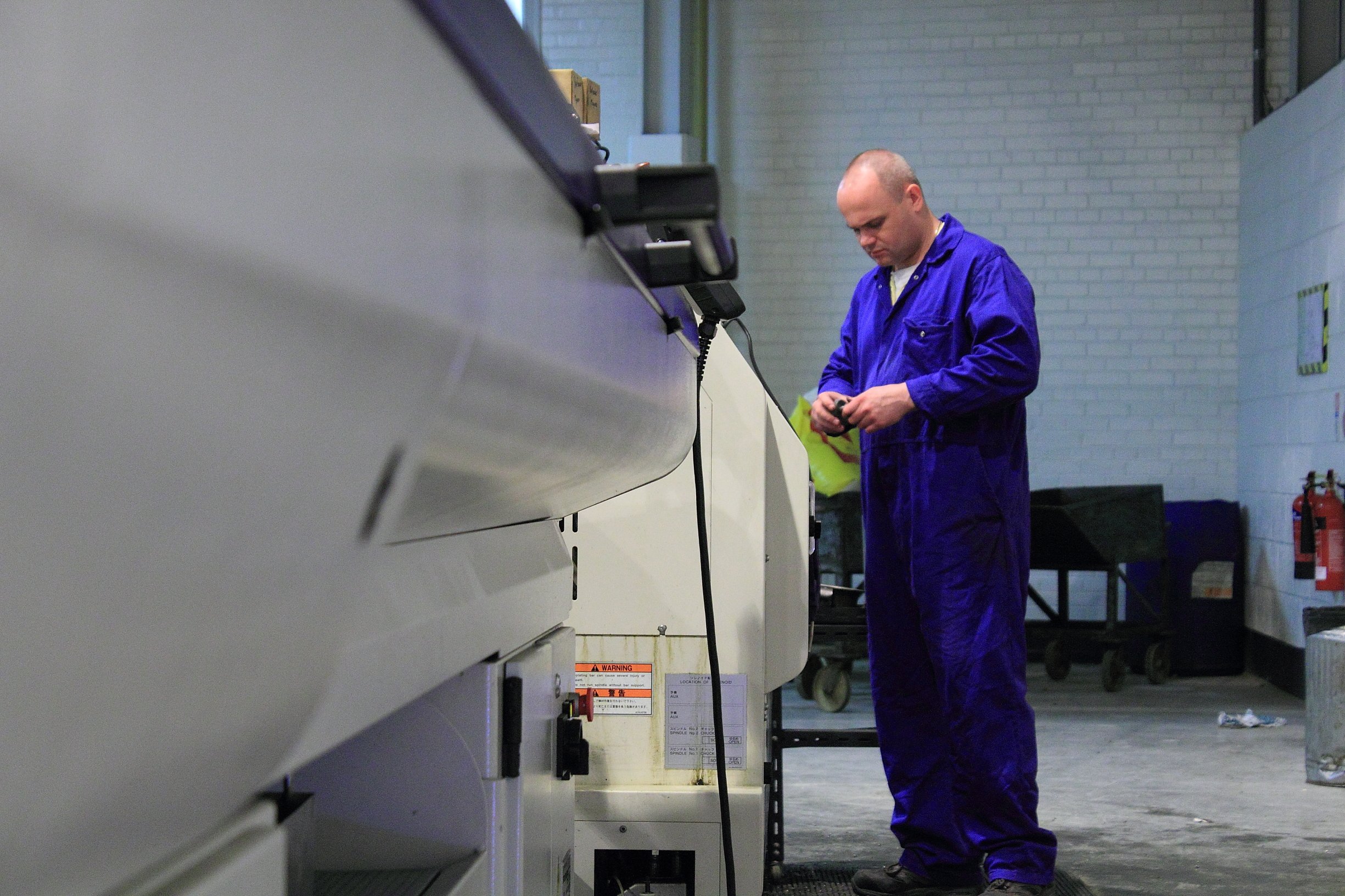 A Factory Worker In Blue Inspects A Precision Grease Nipple Beside Industrial Machines, With Tools And Safety Equipment In The Background.